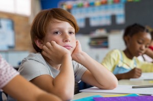 Tired school boy with hand on face sitting at desk in classroom. Bored schoolchild sitting at desk with classmates in classroom. Frustrated and thoughtful young child sitting and looking up.